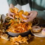Unrecognizable female with tasty unhealthy crisps in hands sitting at table with various junk food in light kitchen at home